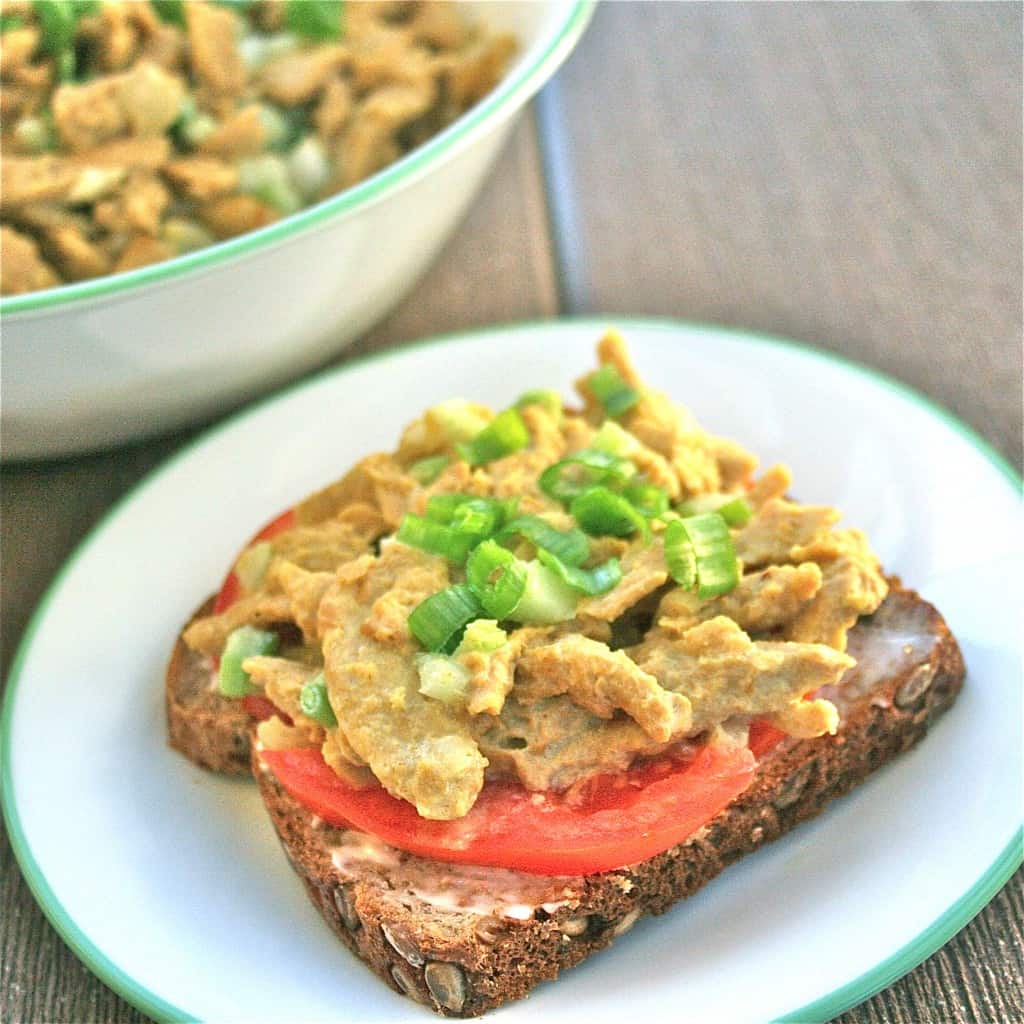 soy curl sandwich spread with slice of tomato on bread, sitting on white plate with wooden background