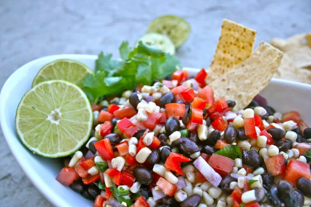 Side view of southwest black bean salad in a white dish with stone background, garnished with limes, cilantro, and chips.