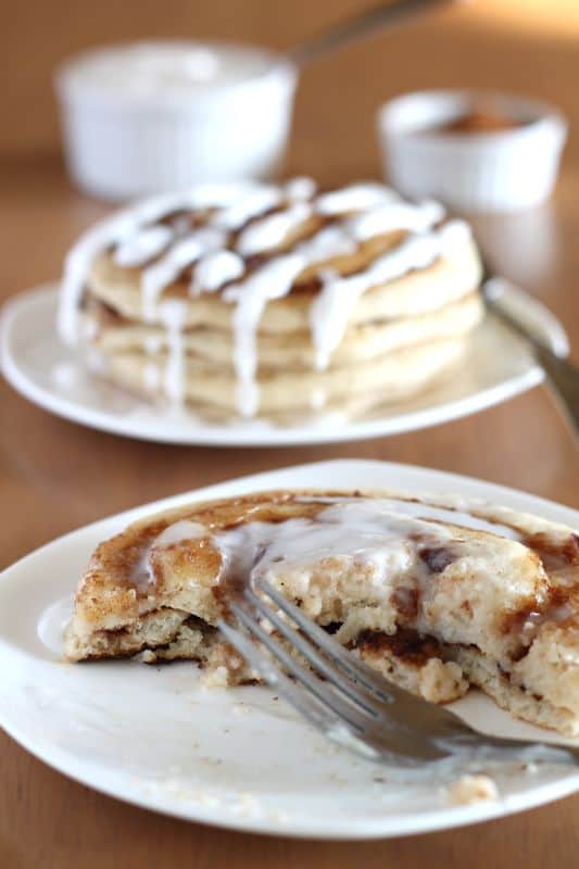 fork with partially eaten vegan cinnamon roll pancakes on white plate with coconut cream topping and wood background