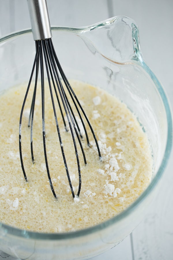 clear glass mixer bowl filled with unmixed vegan lemon cake batter with black whisk