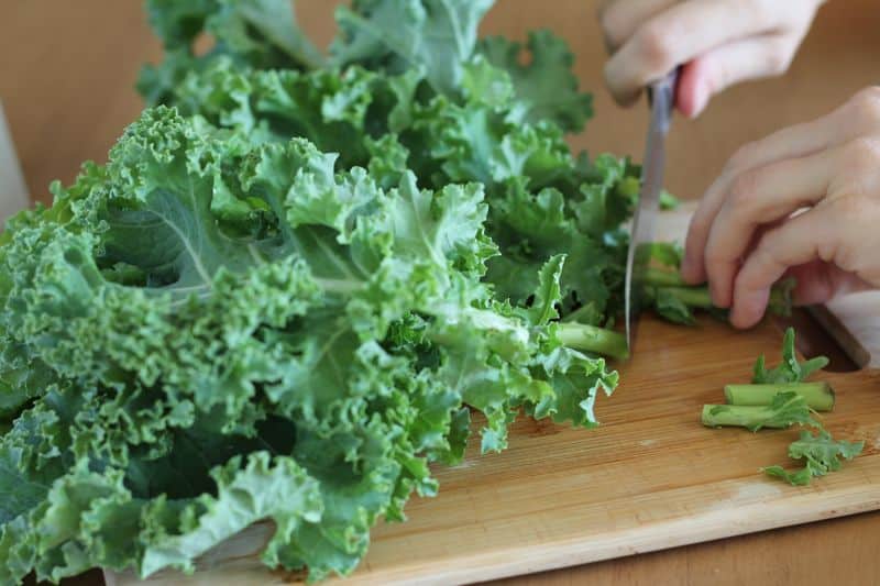 hands chopping lower tough stems from kale leaves on wood cutting board