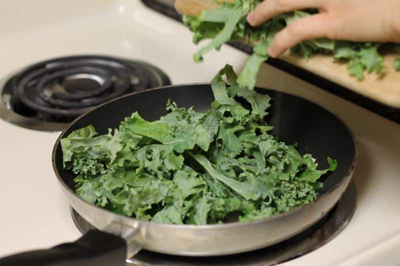 uncooked kale being placed in skillet on stovetop