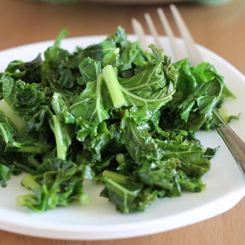 square image of garlic cooked kale on white plate with fork and wooden background