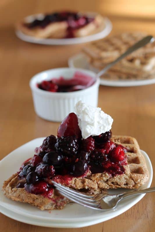 healthy vegan waffles with berry compote and coconut cream on white plate with fork and wood background