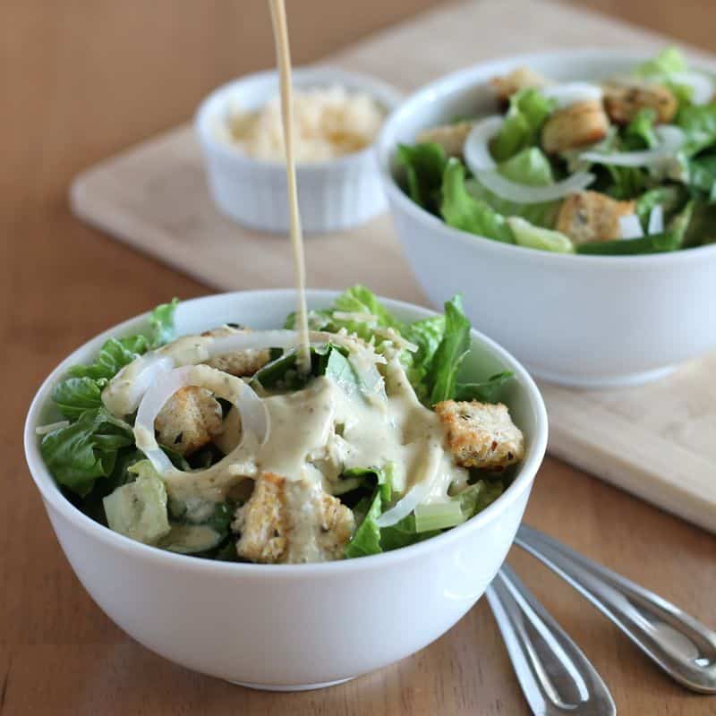vegan Caesar salad dressing being poured over salad in white bowl with wood background