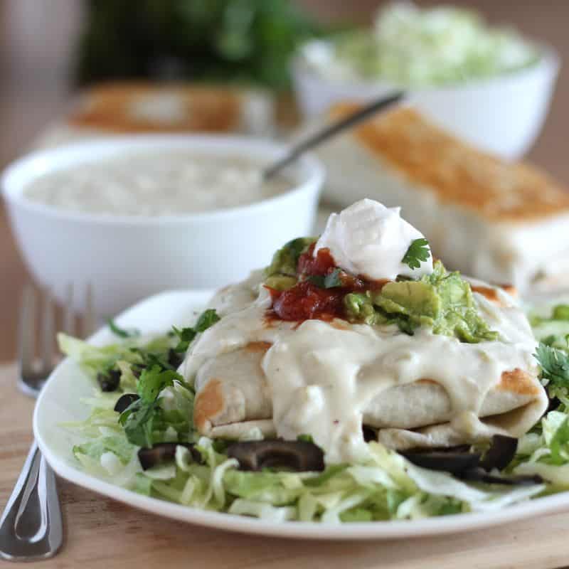 finished chimichanga on white plate with bed of lettuce, and cashew cream, guacamole, salsa, and sour cream toppings