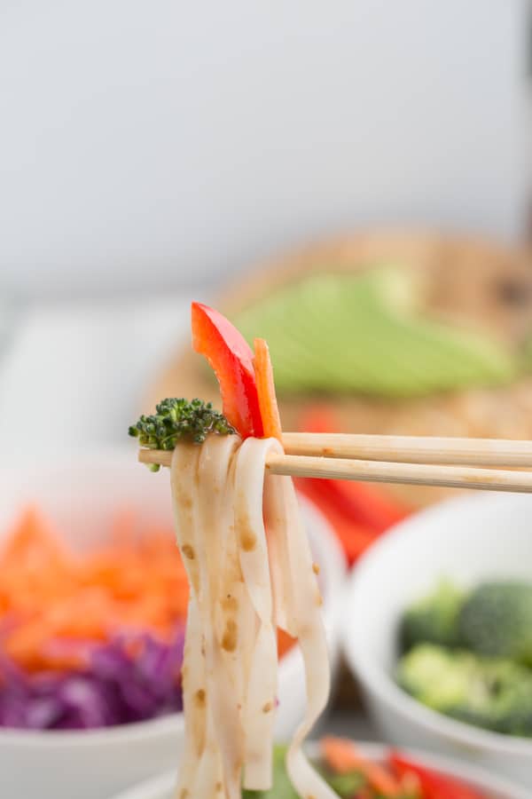 rice noodles with broccoli, pepper, and carrot held between 2 chopsticks, dripping with peanut sauce