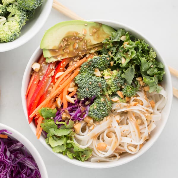 colorful vegetables with rice noodles and peanut sauce arranged in a white bowl with marble background