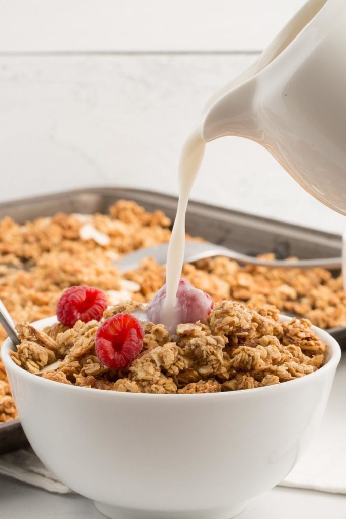 vegan granola in white bowl with nondairy milk being poured into it with white shiplap background
