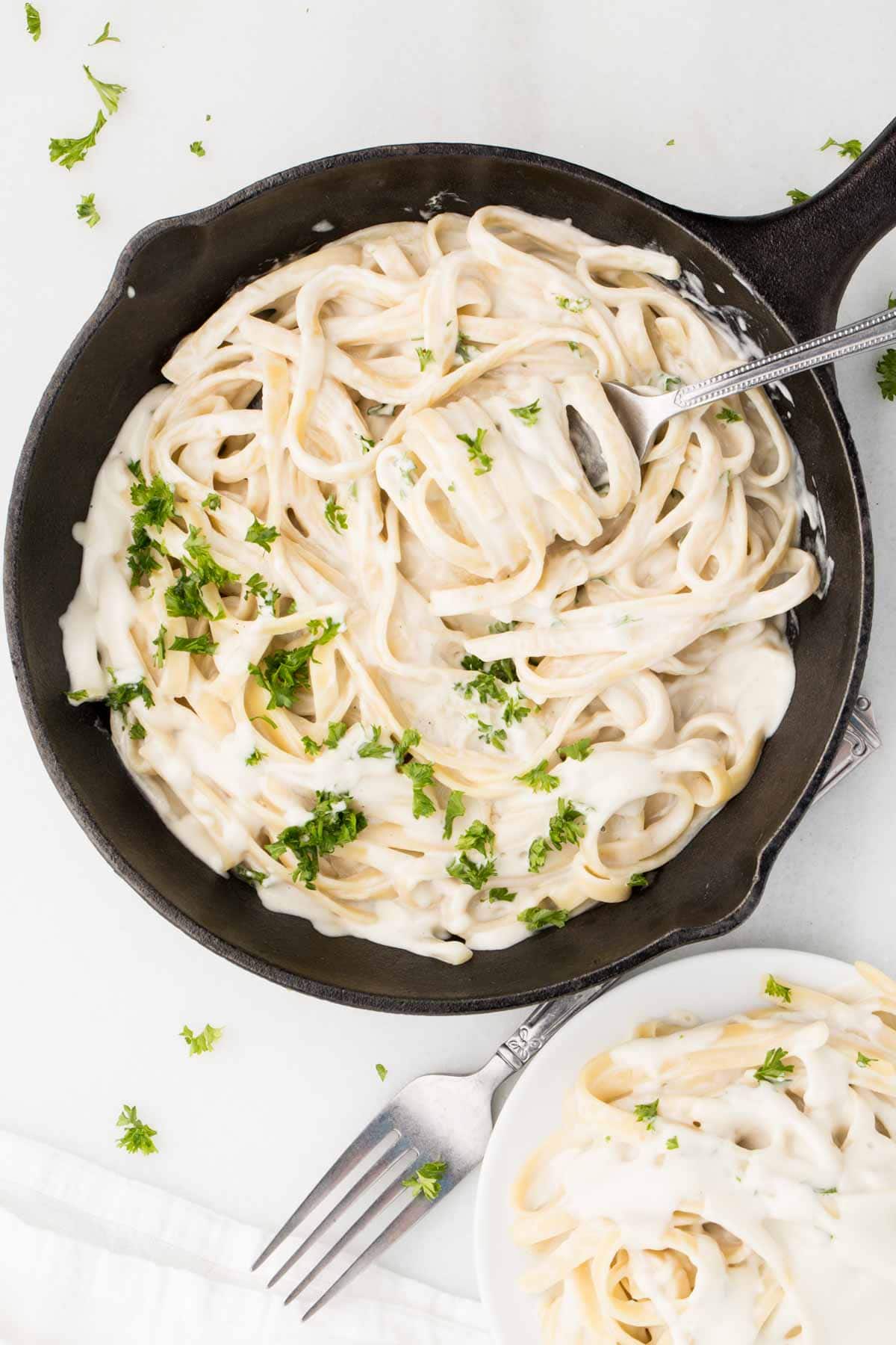 cast iron skillet full of vegan fettuccine alfredo, garnished with parsley, small white plate in right bottom corner of image