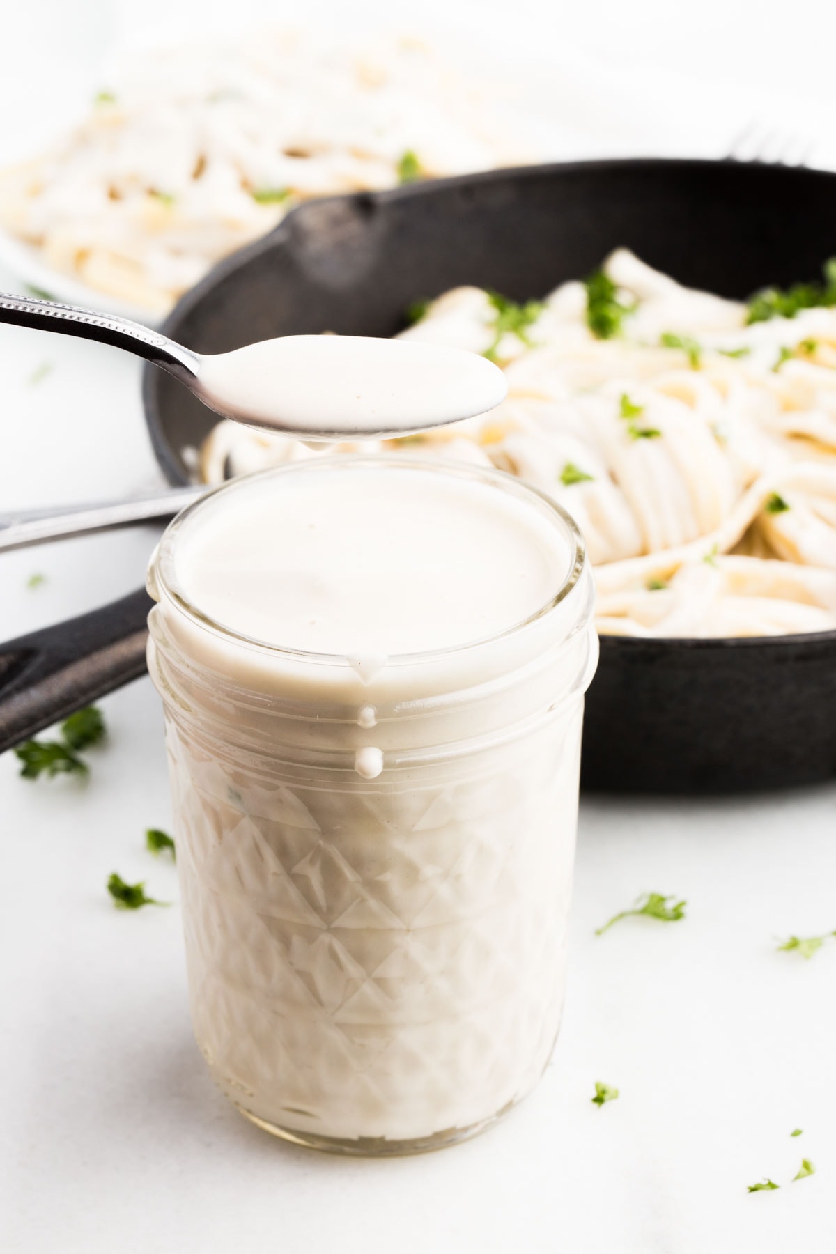 vegan alfredo sauce in spoon held over clear glass jar with cast iron skillet in background