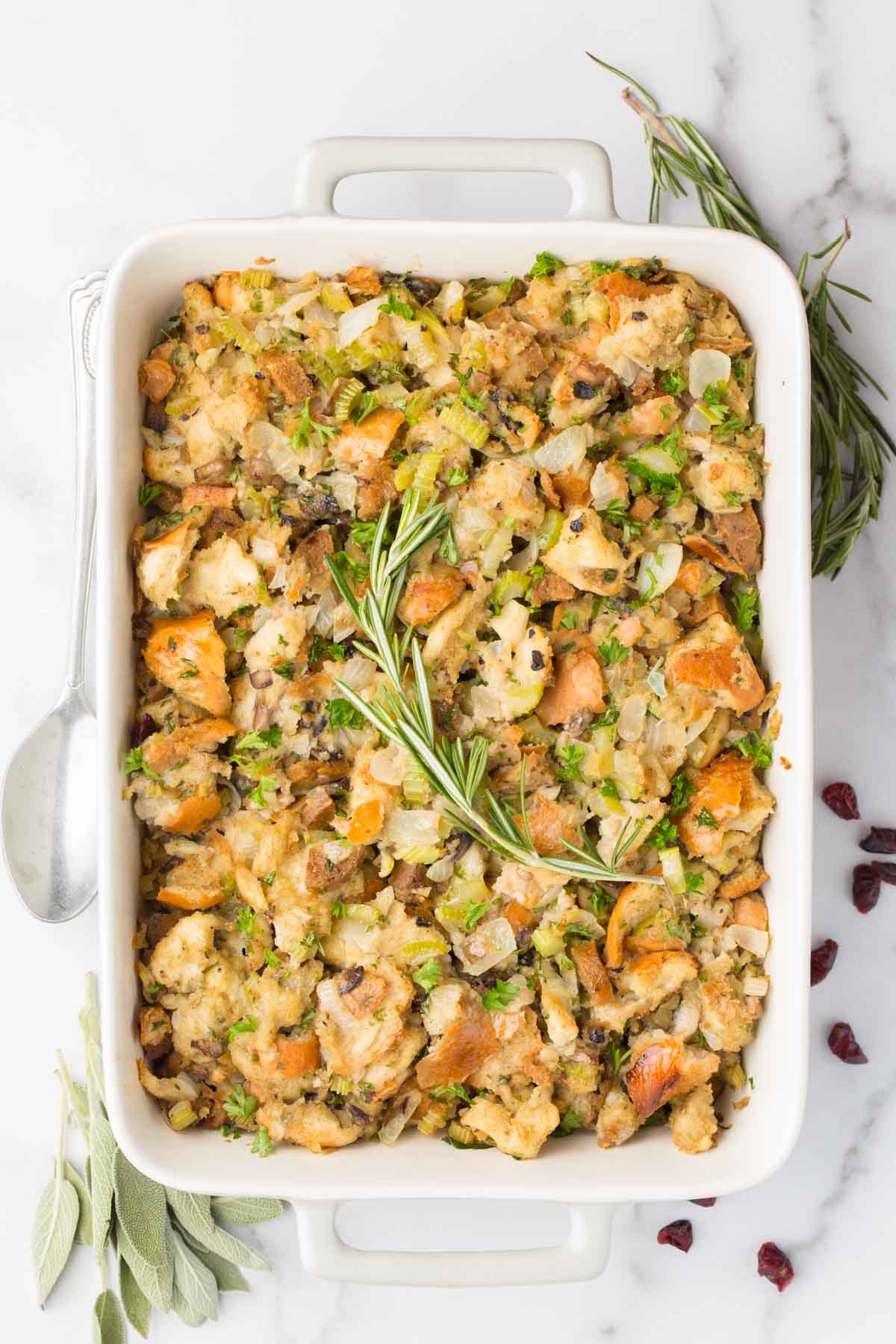 overhead shot of cooked vegan stuffing in white and gray baking dish with spoon on the side and white marble background. Garnished with dried cranberries